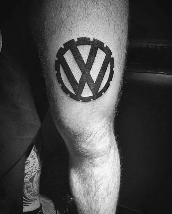 Bold black Volkswagen logo tattoo with gear-like edges, placed on the back of the leg just above the knee.