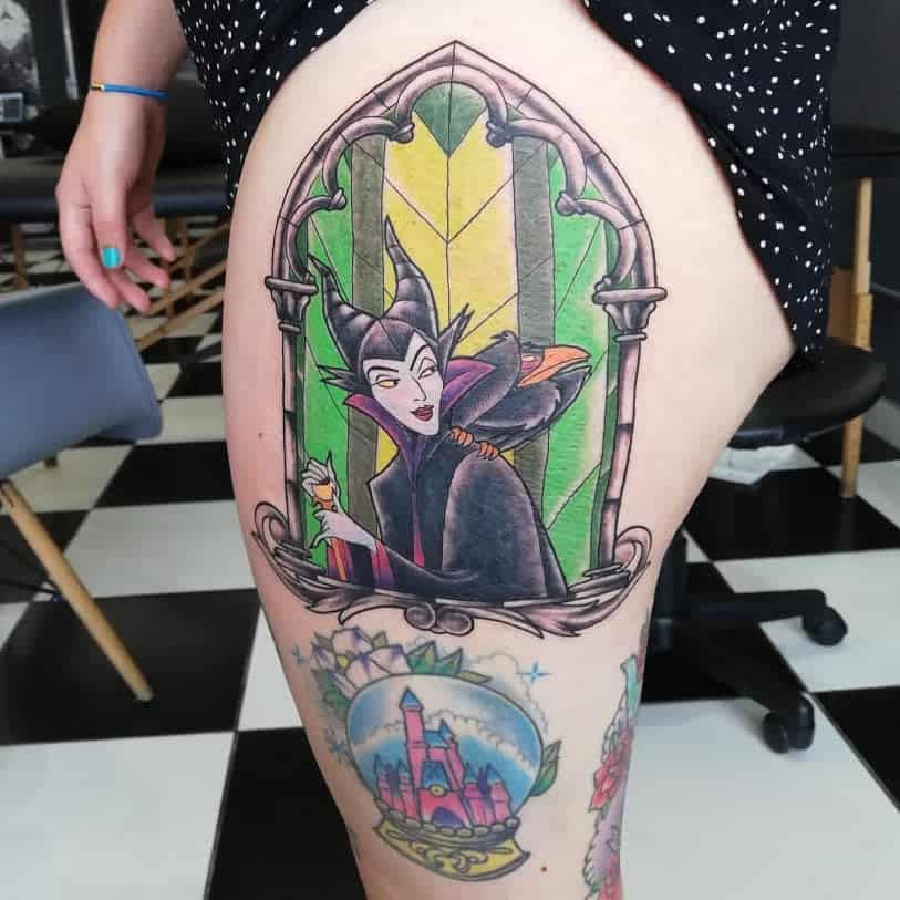 A colorful tattoo of Maleficent as a dark sorceress on a thigh, next to a castle tattoo