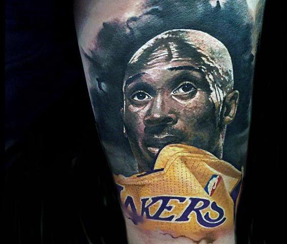 A detailed tattoo of Kobe Bryant, looking upwards with sweat on his face, as he pulls his Lakers jersey over his head, capturing his intense determination.