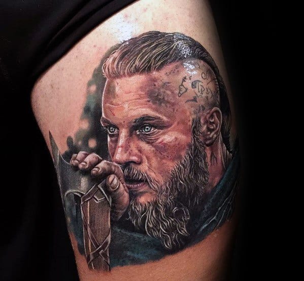 Detailed color tattoo of Ragnar Lothbrok with intense blue eyes, holding a sword hilt.