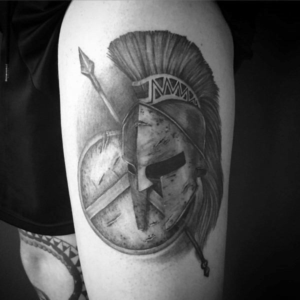 Black and grey upper arm tattoo of a detailed Spartan helmet with a crest, accompanied by a round shield and spear, symbolizing warrior spirit.