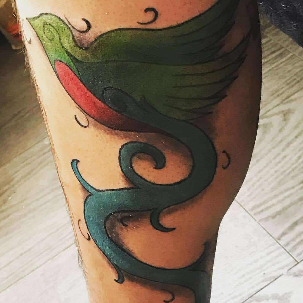 Stylized quetzal tattoo on the thigh, featuring a minimalist green and red bird with long, flowing, swirling tail feathers in a bold design