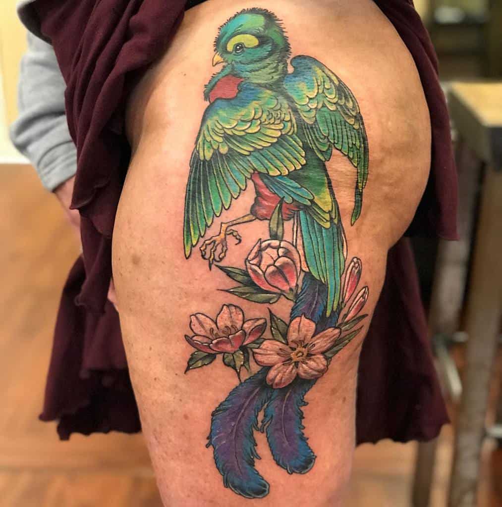 Large, colorful quetzal tattoo on the thigh, featuring a vibrant green and red bird perched among detailed pink flowers with flowing tail feathers