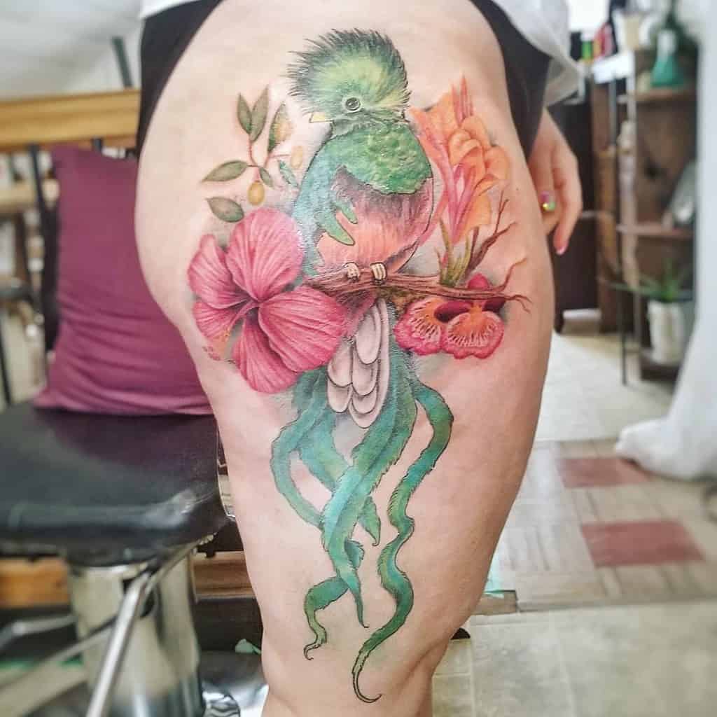 Large and detailed quetzal tattoo on the thigh, featuring a vibrant green and red bird perched on a branch, surrounded by lush pink and orange flowers with flowing tail feathers