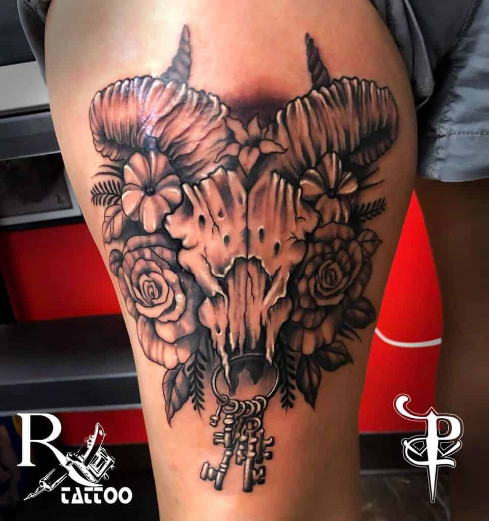 Tattoo of a beautifully detailed ram skull adorned with flowers and keys on a person's upper thigh