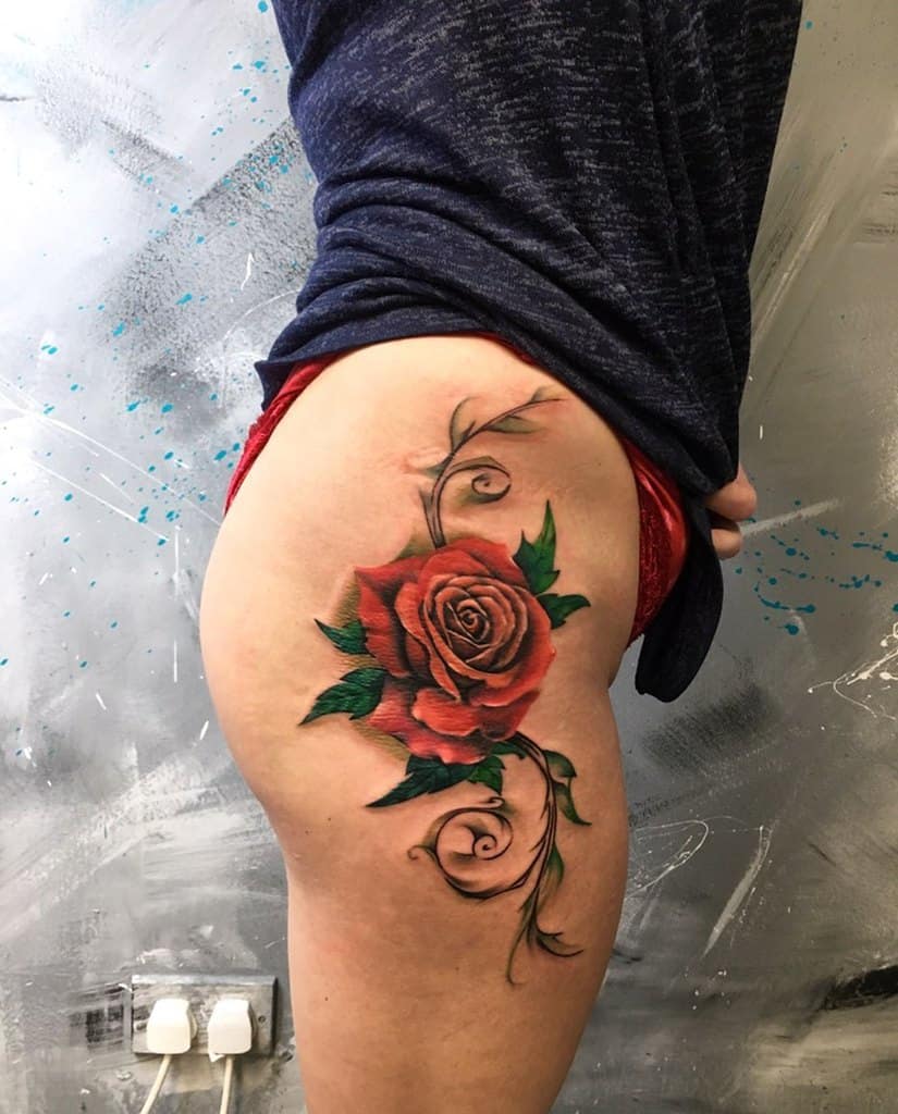 Red rose tattoo on a person's thigh with green leaves and swirling vines
