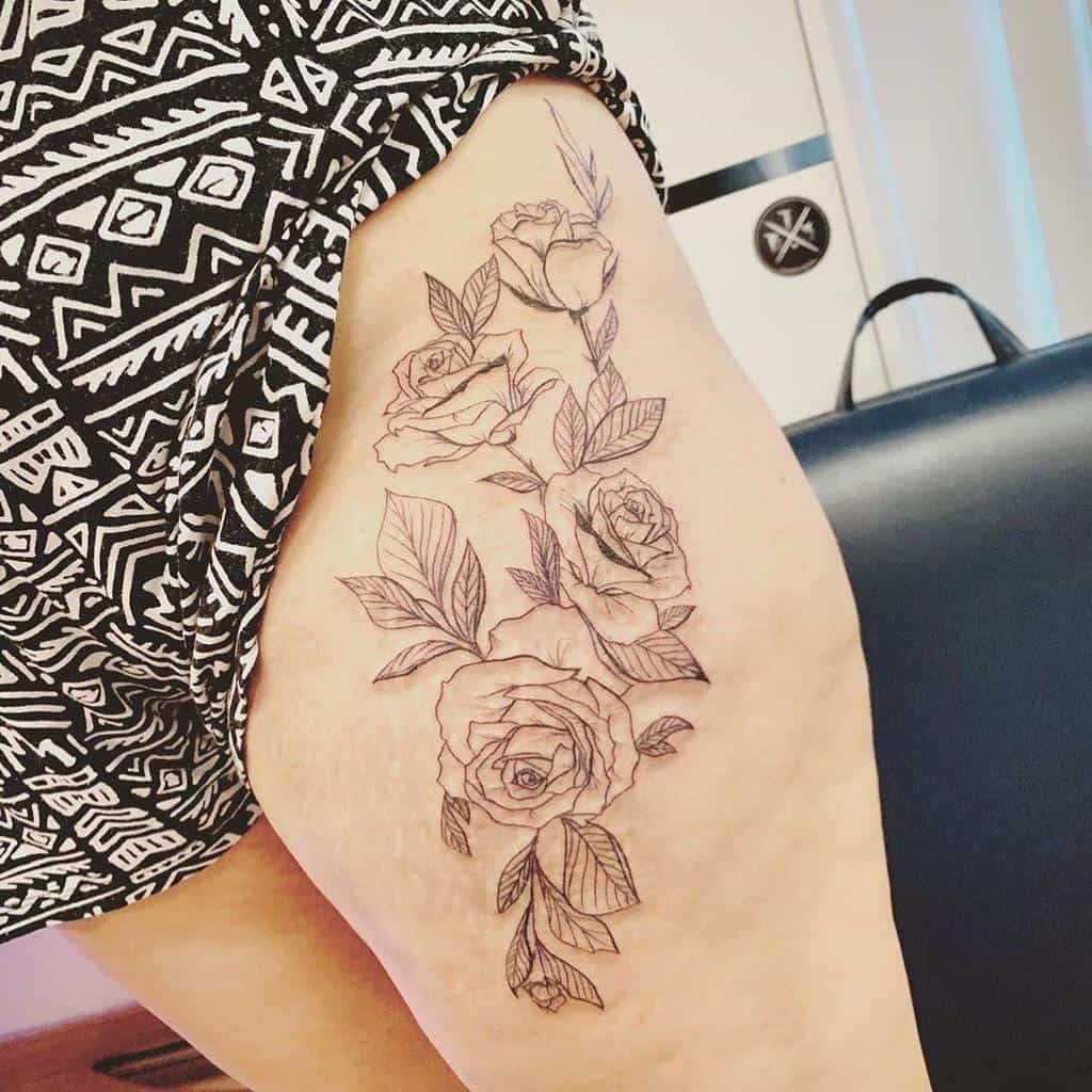 This thigh tattoo features a minimalist design with finely detailed roses and leaves arranged vertically, emphasizing an elegant and natural flow across the leg