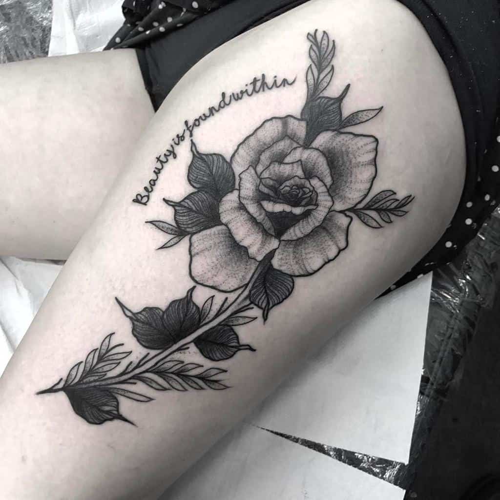 Intricate black and gray thigh tattoo of a blooming rose with detailed leaves and the phrase 'Beauty is found within'
