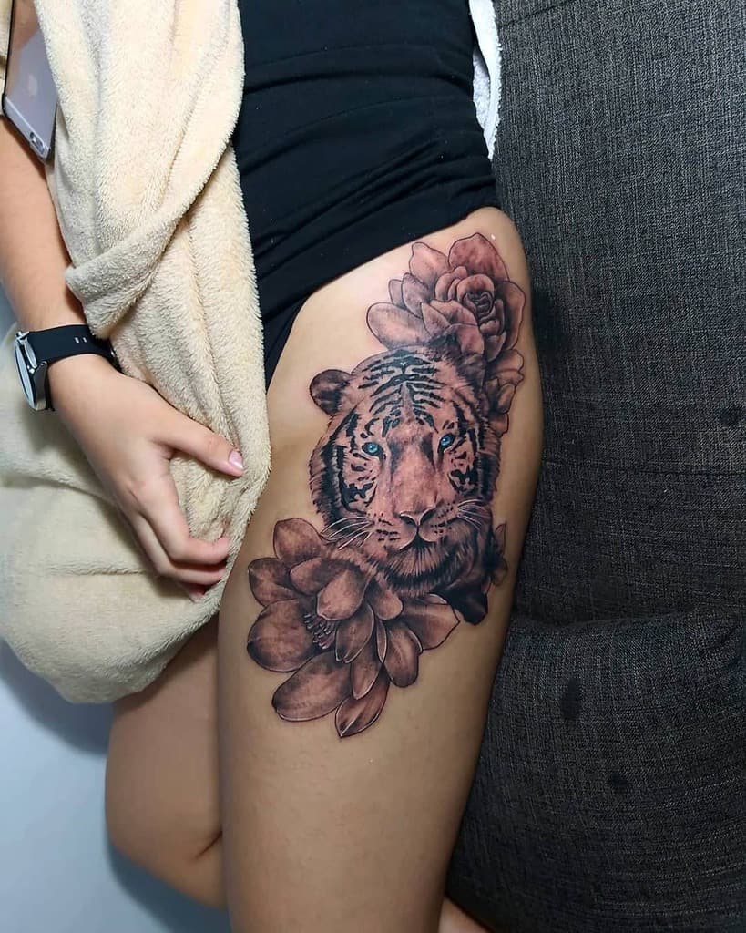 A striking thigh tattoo features a majestic tiger stealthily emerging from a lush bed of flowers on a leg
