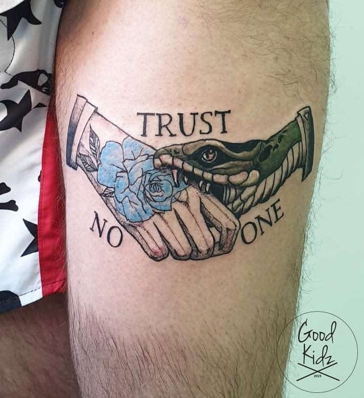 Tattoo of a snake and handshake with blue flower, text "Trust No One" on a person's thigh