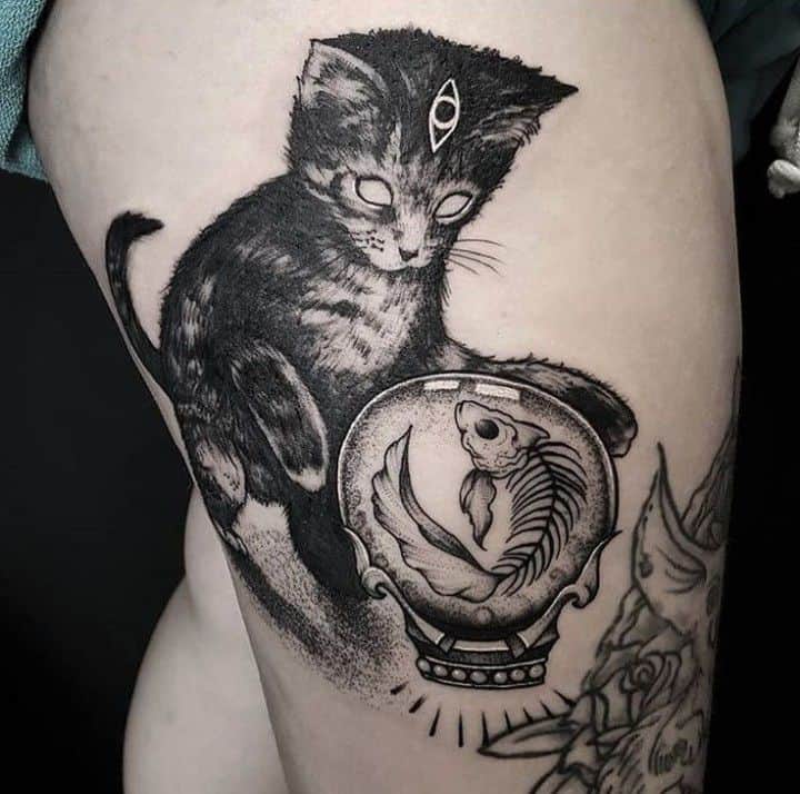 Tattoo of a black cat with a third eye staring into a crystal ball containing a skeletal fish, inked in bold black shading on the thigh.