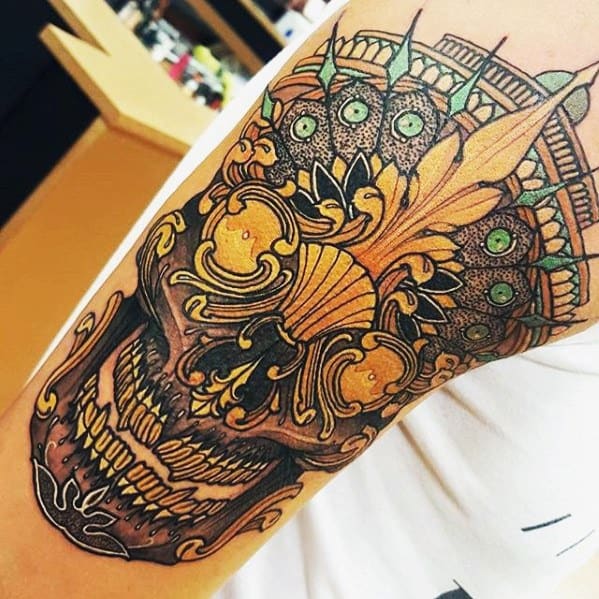 Tibetan skull tattoo on the upper arm with intricate golden detailing, green gem accents, and bold ornamental crown design.