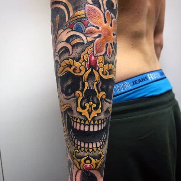 Tibetan skull tattoo on the forearm with golden ornamental crown, wide grin, and vibrant orange flower at the top.