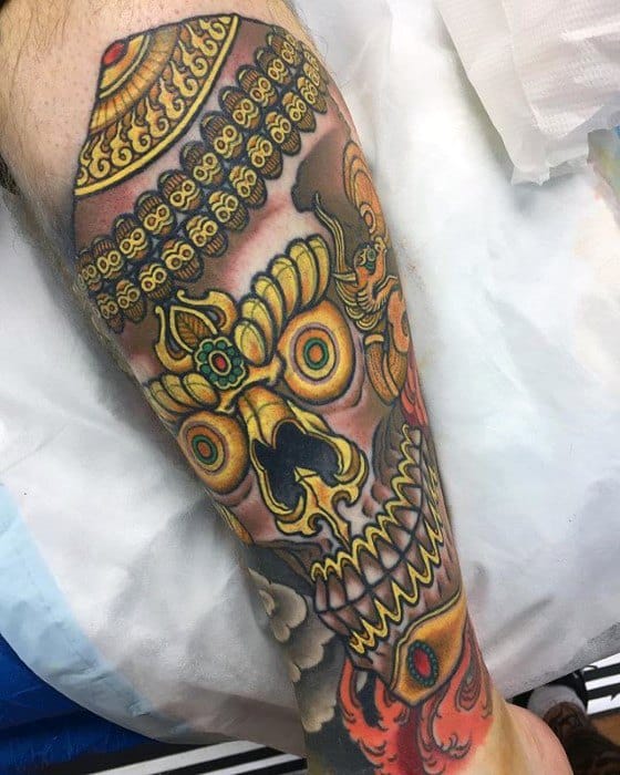 Colorful Tibetan skull tattoo on the leg with intricate golden crown, red jewel accents, and bold teeth outlined in deep black.