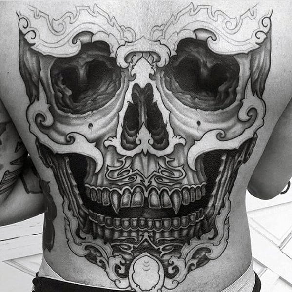 Black and grey Tibetan skull tattoo covering the full back with deep-set eyes, sharp teeth, and swirling ornamental patterns.