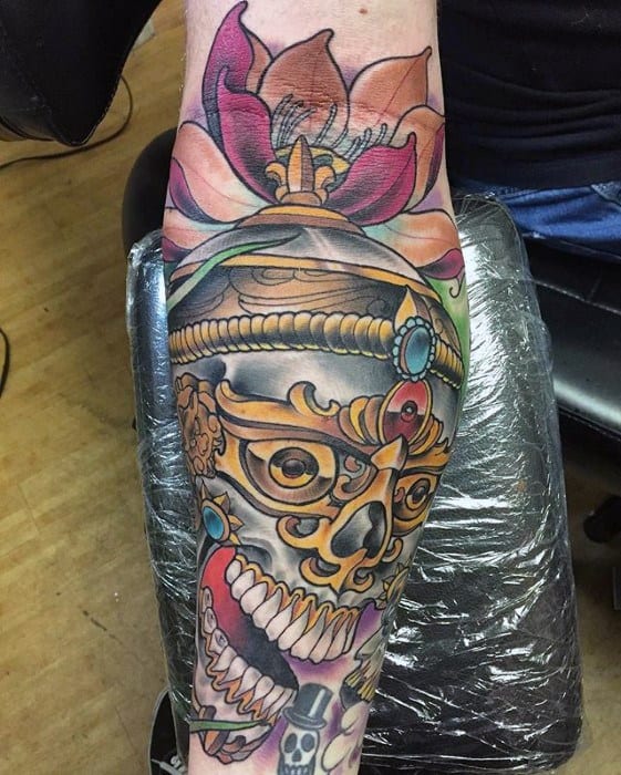 Colorful Tibetan skull tattoo on the leg with jeweled crown, lotus flower on top, and bold teeth design stretching from ankle to knee.