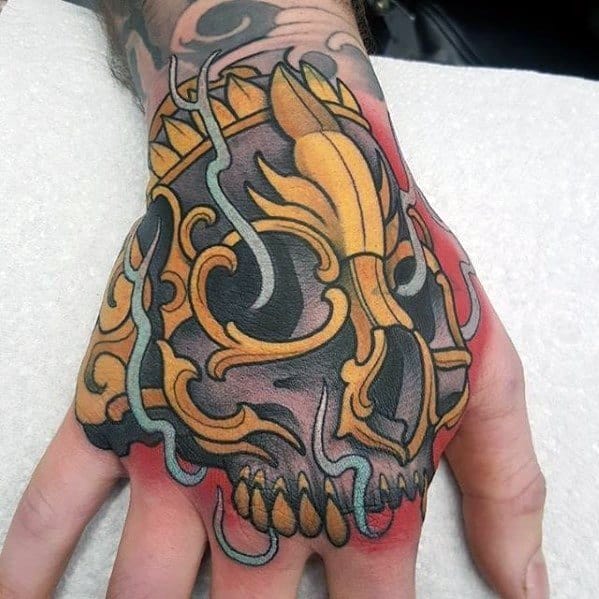 Tibetan skull tattoo on the hand with bold black outlines, golden ornamental patterns, and red shading for a fiery effect.