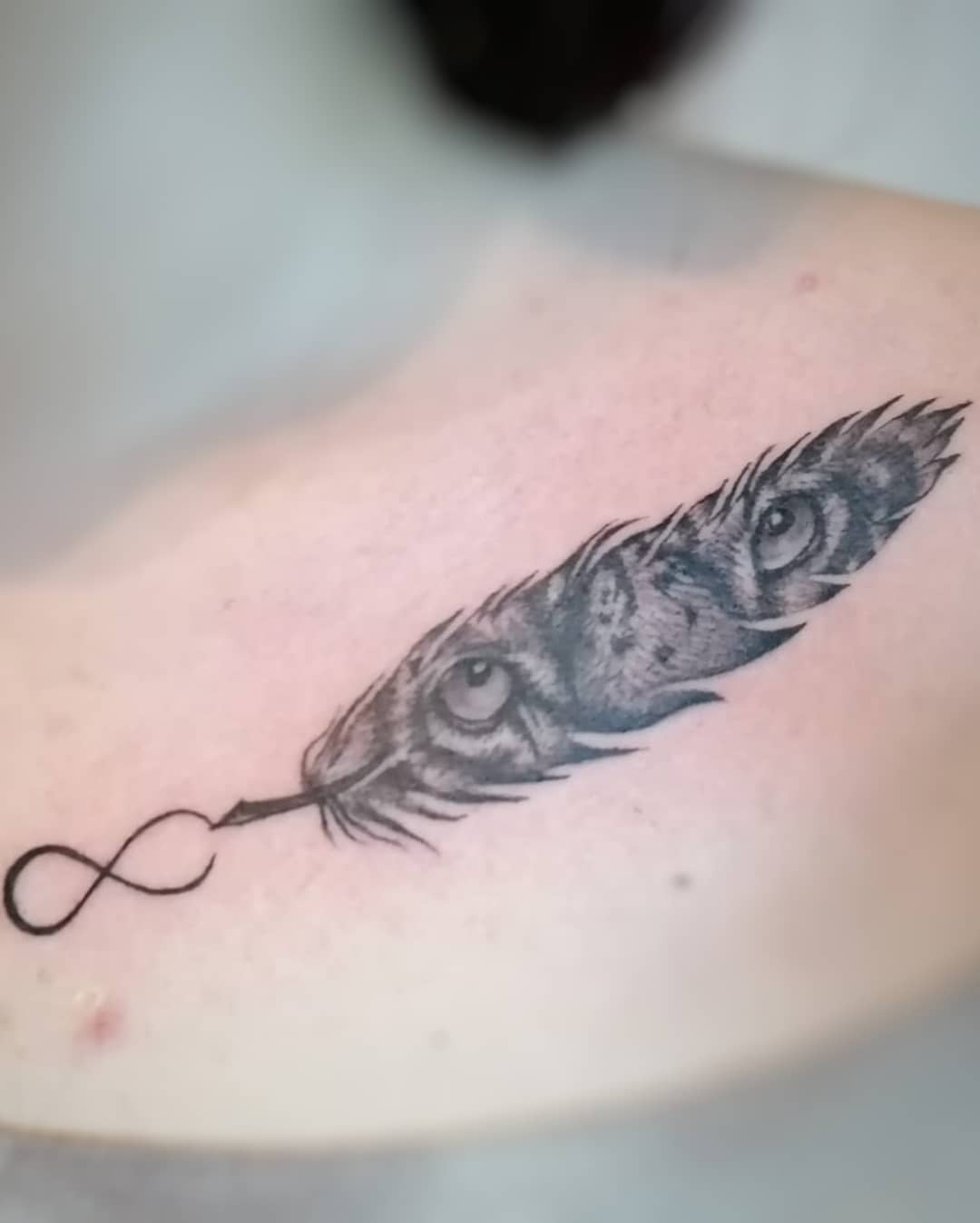 Tattoo of a feather with tiger eyes and an infinity symbol on a shoulder