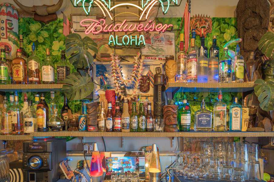 Colorful tiki bar with tropical decor, neon "Aloha" sign, and an assortment of liquor bottles and glasses.