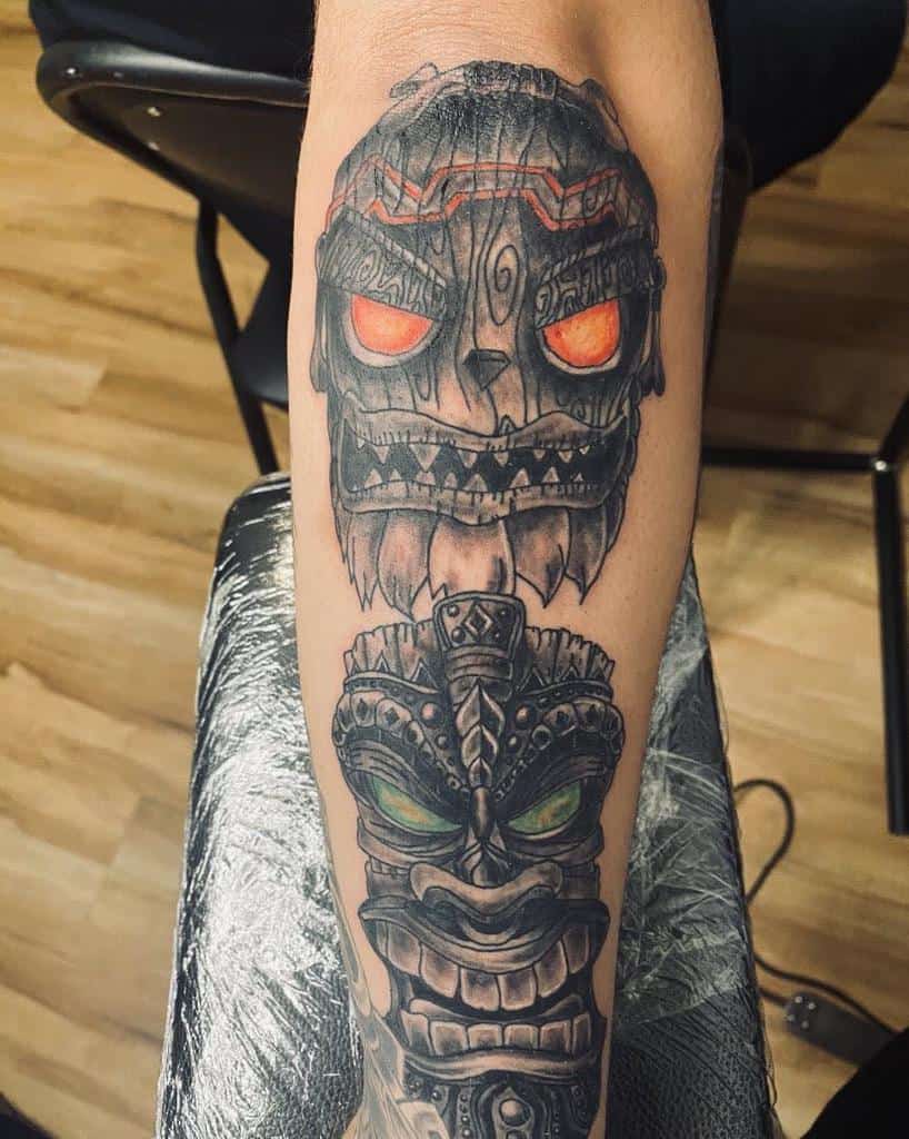 A black-and-gray tattoo of two Tiki masks, one with glowing orange eyes and sharp teeth, and the other with green eyes