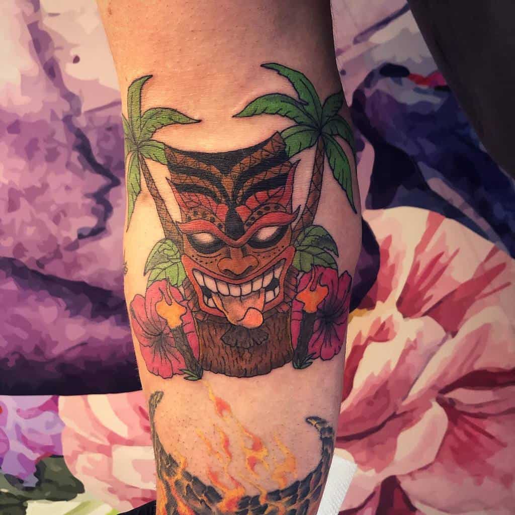 A vibrant tattoo of a Tiki mask surrounded by palm trees and hibiscus flowers, featuring bold colors and lively details
