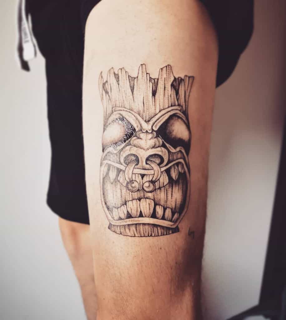A black-and-gray tattoo of a Tiki mask carved from wood, featuring sharp teeth and a fierce expression