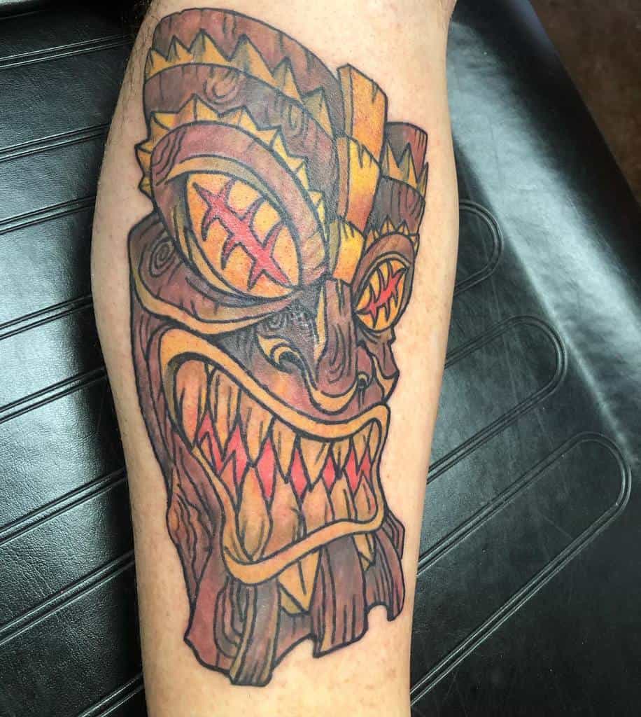 A vibrant, colorful tattoo of a Tiki mask, featuring sharp teeth, intricate geometric patterns, and bright shades of orange and yellow