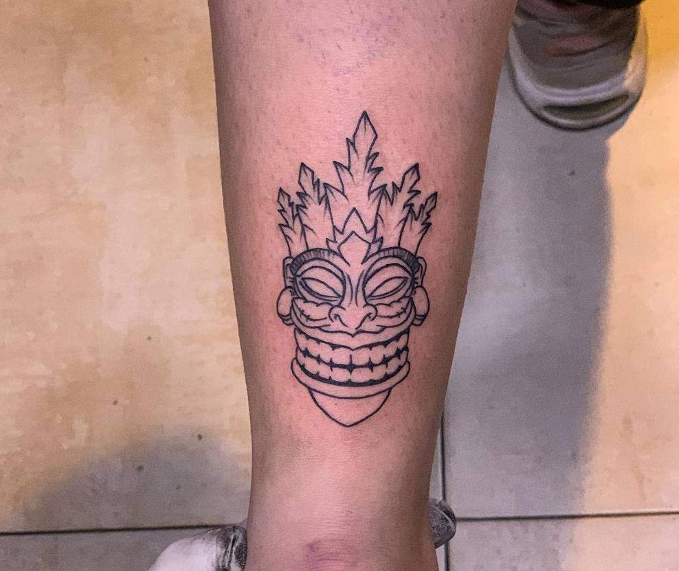 A minimalist black-line tattoo of a Tiki mask, featuring sharp angles and a stylized design with a crown-like top