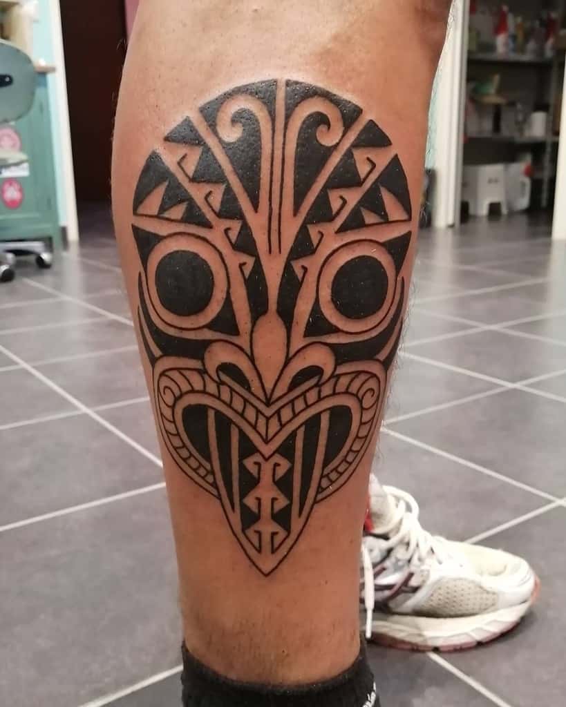 A bold black-and-gray tattoo of a Tiki mask with geometric patterns and swirling motifs