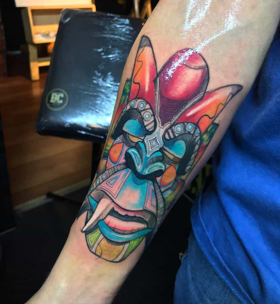 A vibrant, colorful tattoo of a Tiki mask featuring bold blues, reds, and yellows, with intricate patterns