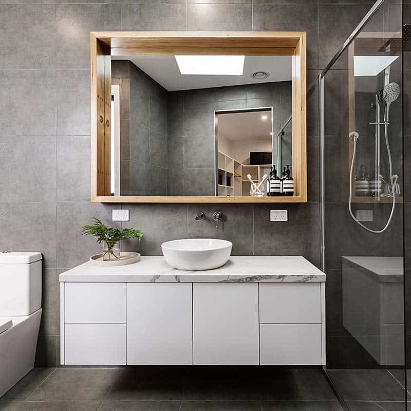 This modern bathroom has grey tiles, a wooden-framed mirror, and a white floating vanity with a glass shower.
