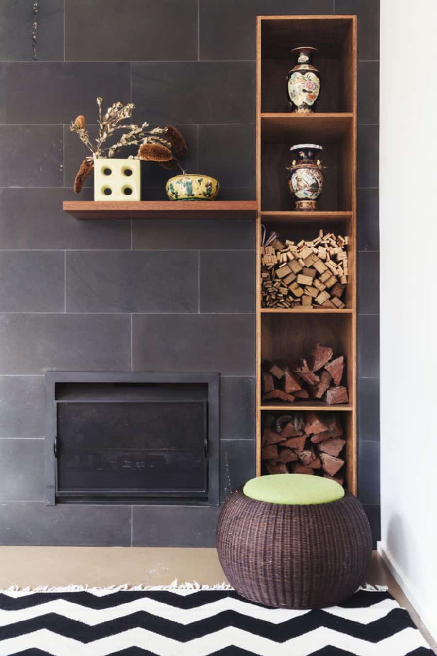 Modern fireplace with decorative shelves, vases, firewood, and a woven ottoman on a patterned rug against an accent wall
