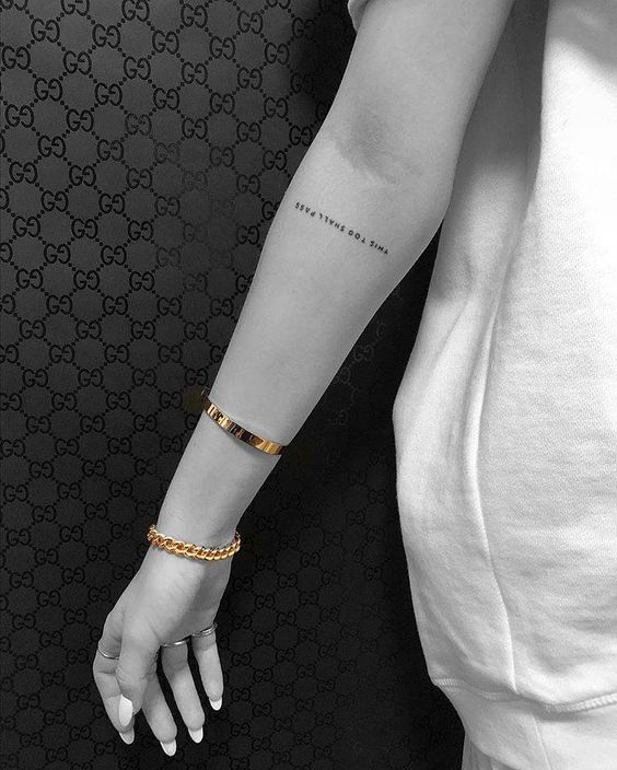 This image shows a tattoo on the arm with the phrase "NEW TIMES GOD BLESS" in a simple, minimalist font. The background features a Gucci pattern.