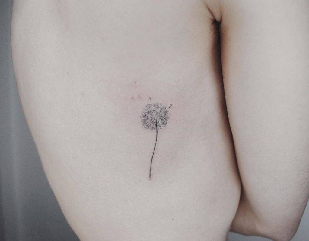 Minimalist dandelion tattoo on the ribcage with delicate seeds drifting away, symbolizing freedom, wishes, and new beginnings.