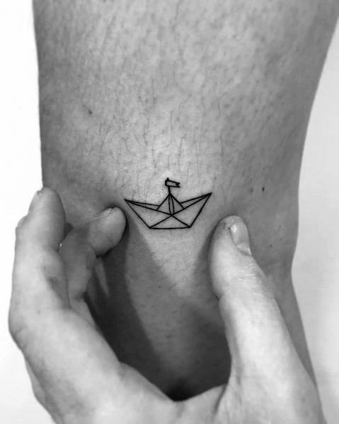 Minimalist black ink tattoo of a small paper boat with a tiny flag, placed on the ankle, symbolizing simplicity, adventure, and resilience.