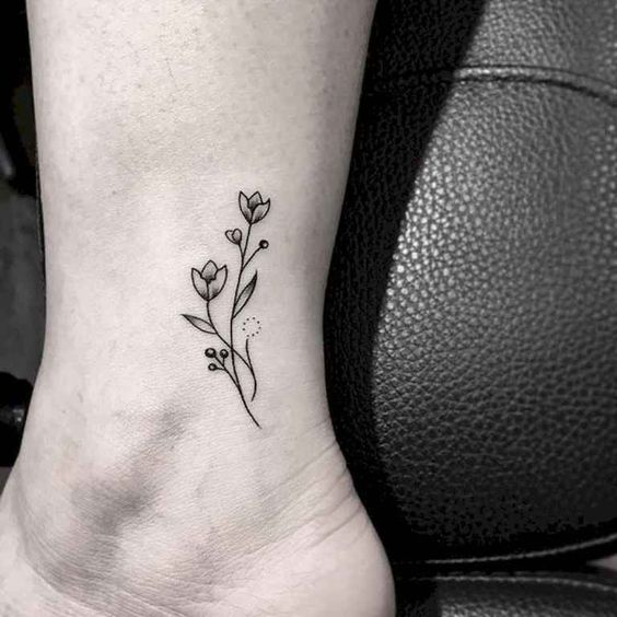 A delicate black-line tulip tattoo on the ankle, featuring two small flowers, leaves, and tiny decorative dots for an elegant minimalist design.