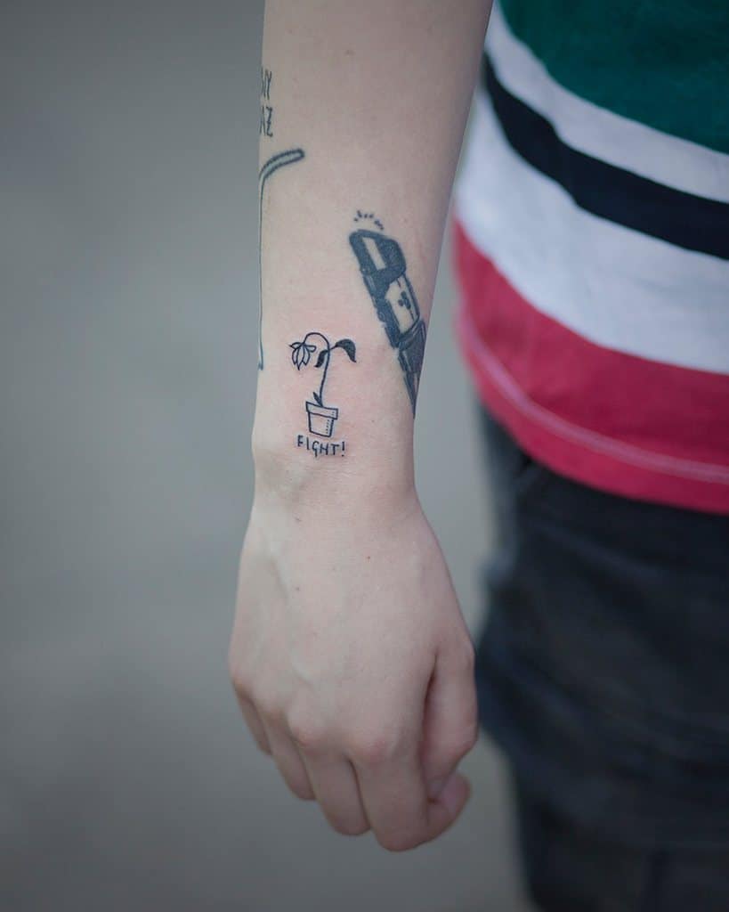 A minimalist black ink tattoo of a small wilted flower in a pot with the word 'FIGHT!' written below, placed on the wrist as a symbolic design.