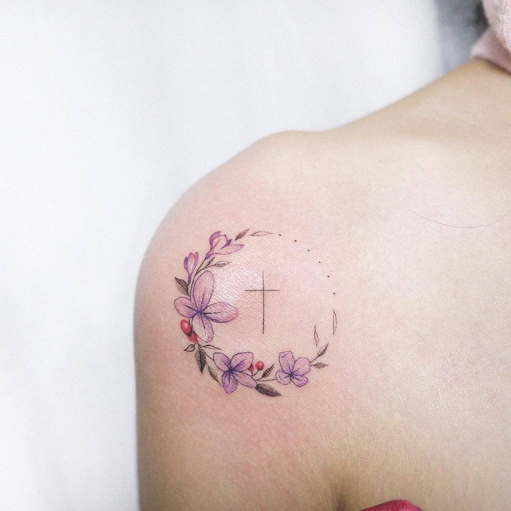 Delicate shoulder tattoo of a floral wreath with soft purple lilac blossoms encircling a minimalist cross, blending fine-line and watercolor styles.