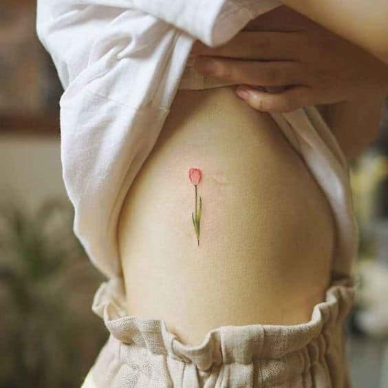 A small, delicate red tulip tattoo on the ribcage, featuring a minimalist design with a thin green stem and subtle shading for a soft, elegant look.