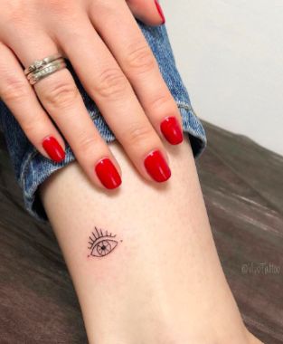 Small minimalist tattoo of an all-seeing eye with radiating lines, inked in black on the ankle.