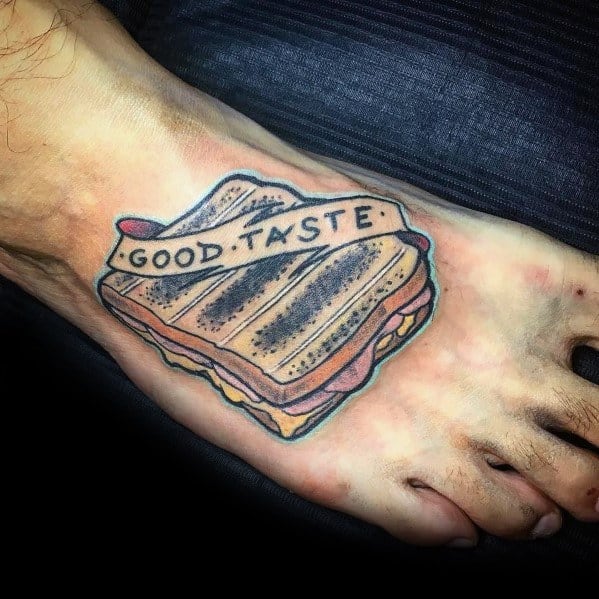 Grilled sandwich tattoo on the foot with a banner reading "Good Taste," featuring bold outlines, shading, and a classic traditional tattoo style.