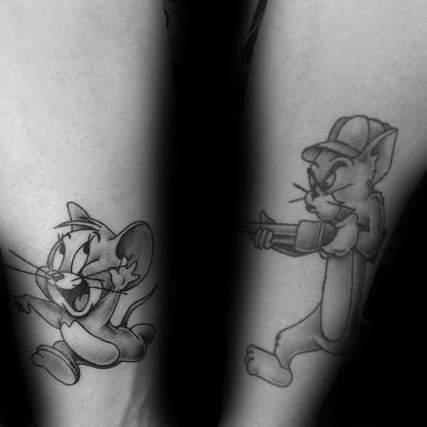 Black and grey matching tattoos of a scared Jerry on one leg and Tom aiming a rifle on the other, in bold cartoon style.