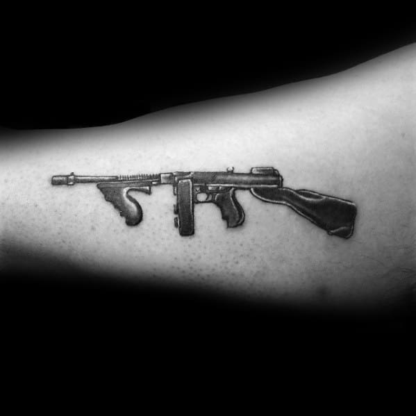 Realistic black and grey Tommy gun tattoo on the forearm, featuring fine shading and intricate details for a bold yet minimal firearm design.