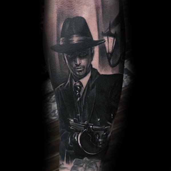 Realistic black and grey gangster tattoo featuring a suited mobster in a fedora holding a Tommy gun, with dramatic shading for a cinematic crime scene look.