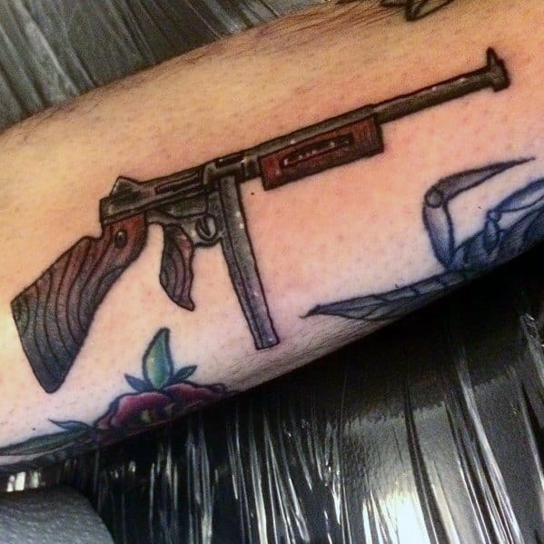 Traditional-style Tommy gun tattoo with bold outlines, wood grain detailing, and subtle shading, creating a vintage firearm design on the forearm.
