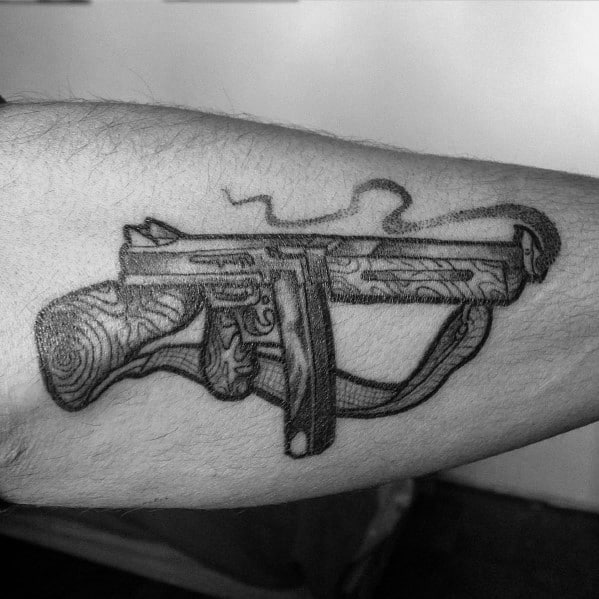 Detailed Tommy gun tattoo with fine linework, wood grain texture, and smoke effects, creating a bold and dynamic vintage firearm design on the forearm.