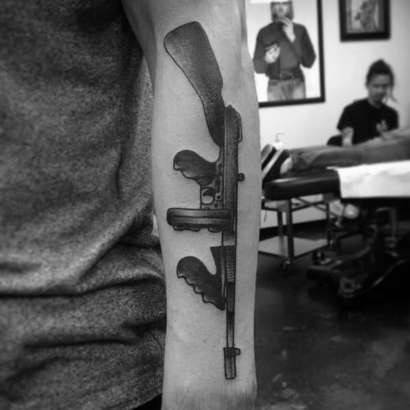 Realistic black and grey Tommy gun tattoo on the forearm, featuring intricate shading and fine details for a bold, vertically positioned firearm design.