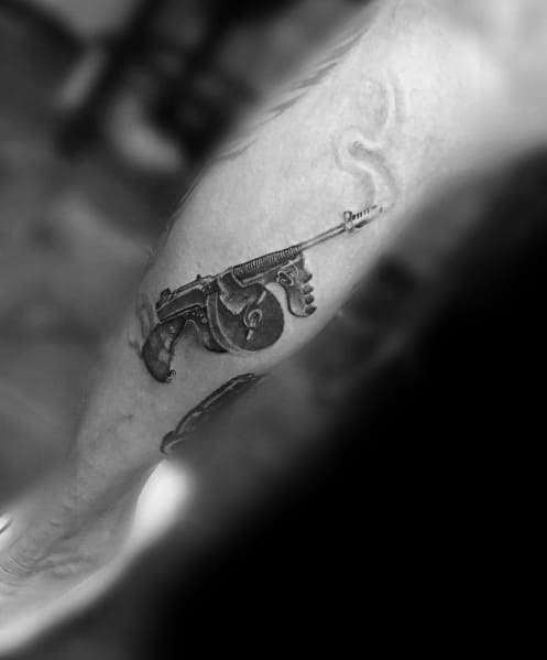 Realistic black and grey Tommy gun tattoo on the forearm, featuring fine details, shading, and subtle smoke effects for a classic vintage firearm look.