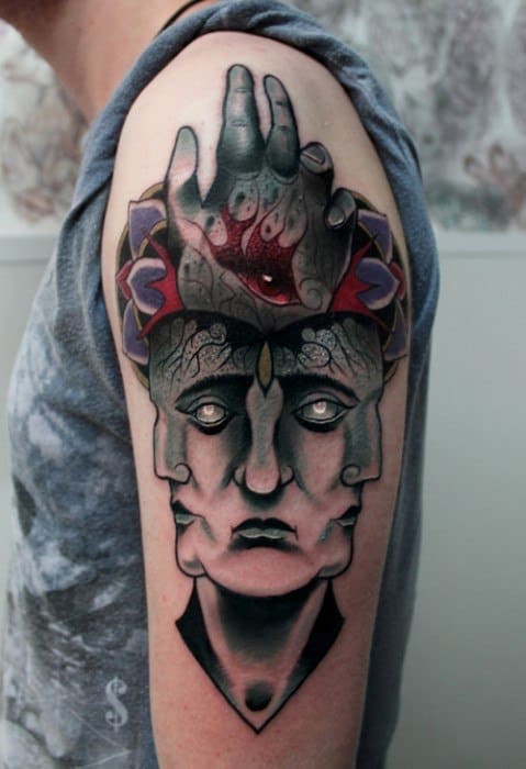 Tool-inspired upper arm tattoo featuring a three-faced surreal figure with a third eye and a stylized hand displaying multiple eyes above it.