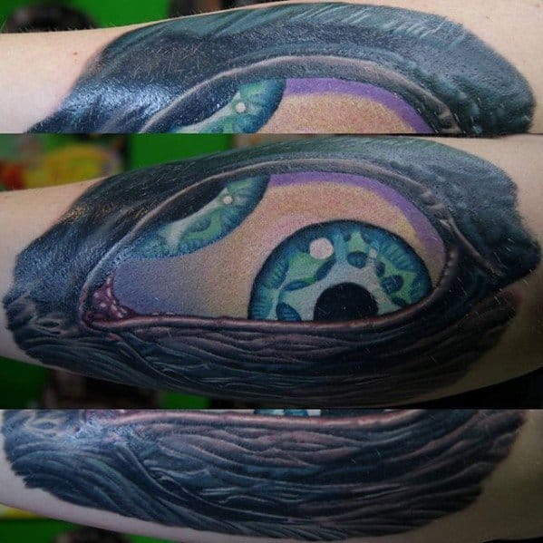 Tool-inspired forearm tattoo showing a vivid blue-green eye peeking through torn skin, with detailed shading and textured 3D realism.